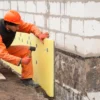 Construction worker in orange safety clothing installing yellow polystyrene insulation boards on an exterior foundation wall with visible mechanical fixings