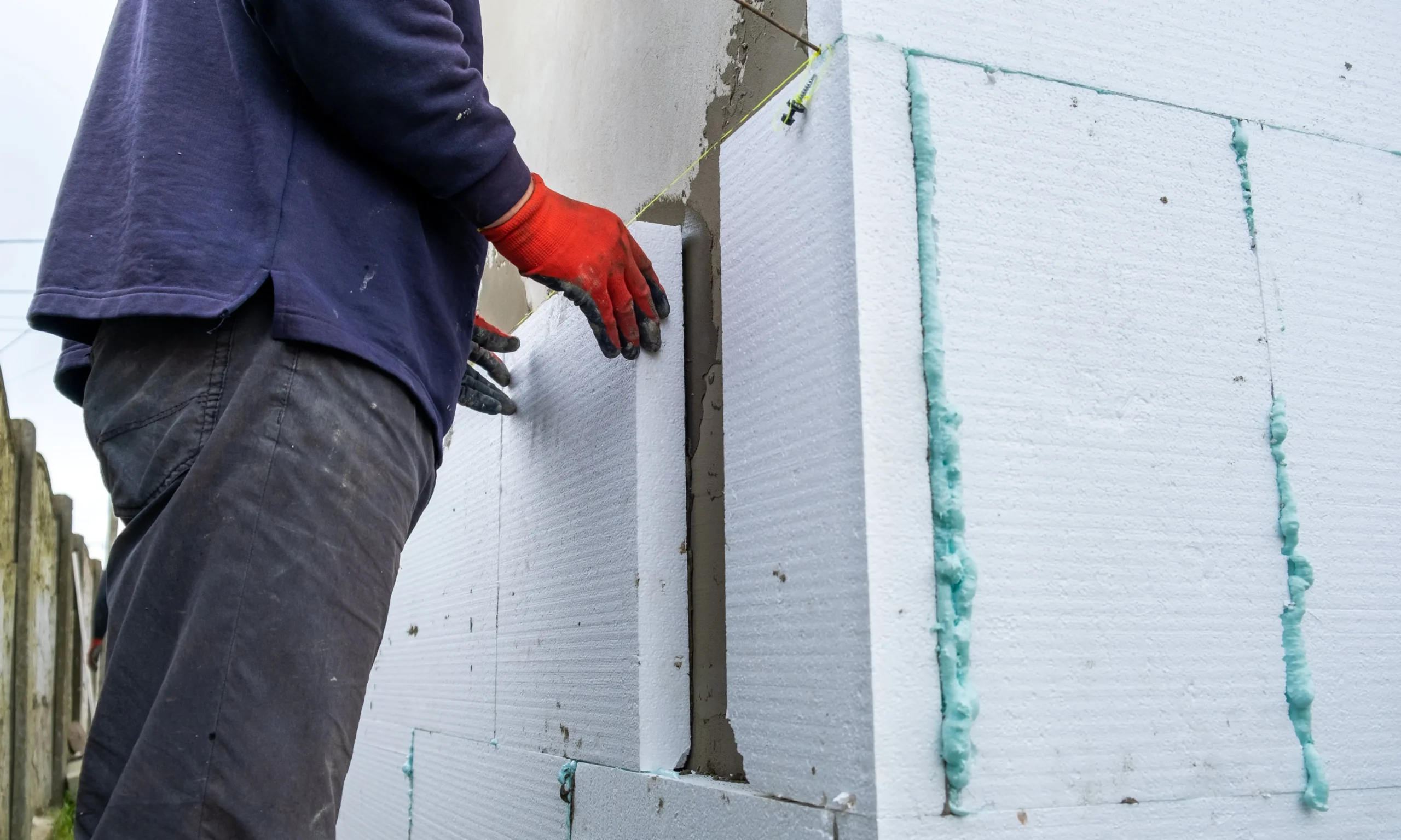 A worker placing polystyrene insulation boards on an exterior wall using adhesive and alignment string