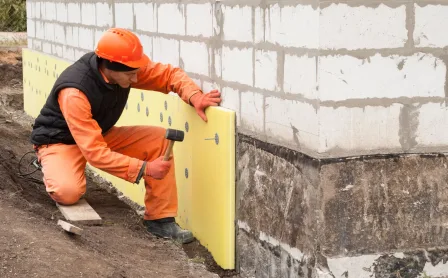 Construction worker in orange safety clothing installing yellow polystyrene insulation boards on an exterior foundation wall with visible mechanical fixings