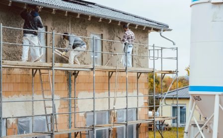 Workers applying cement render to a house wall using tools on a construction site.